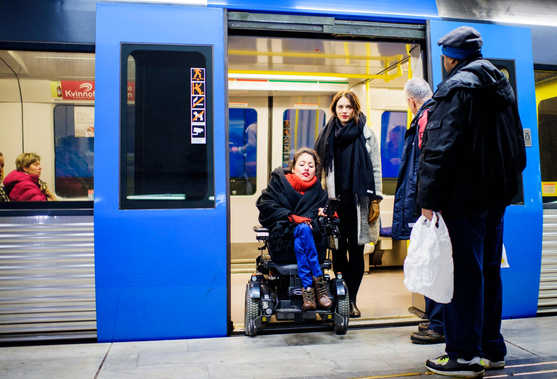 Woman in a wheelchair getting of a train with an assistant. Other people are standing on the platform.