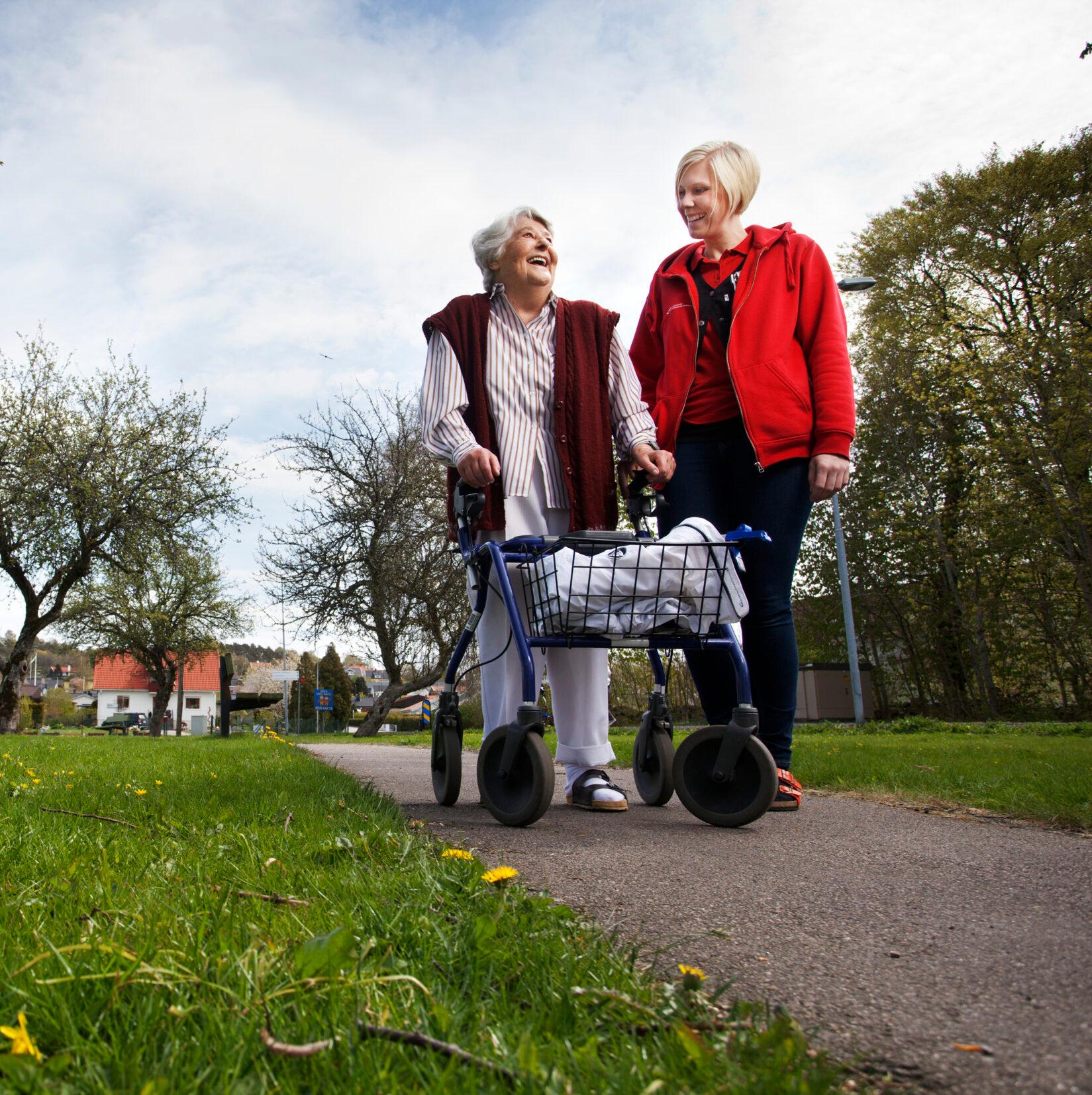 An elderly woman in a park walking with a walking frame together with a younger woman.