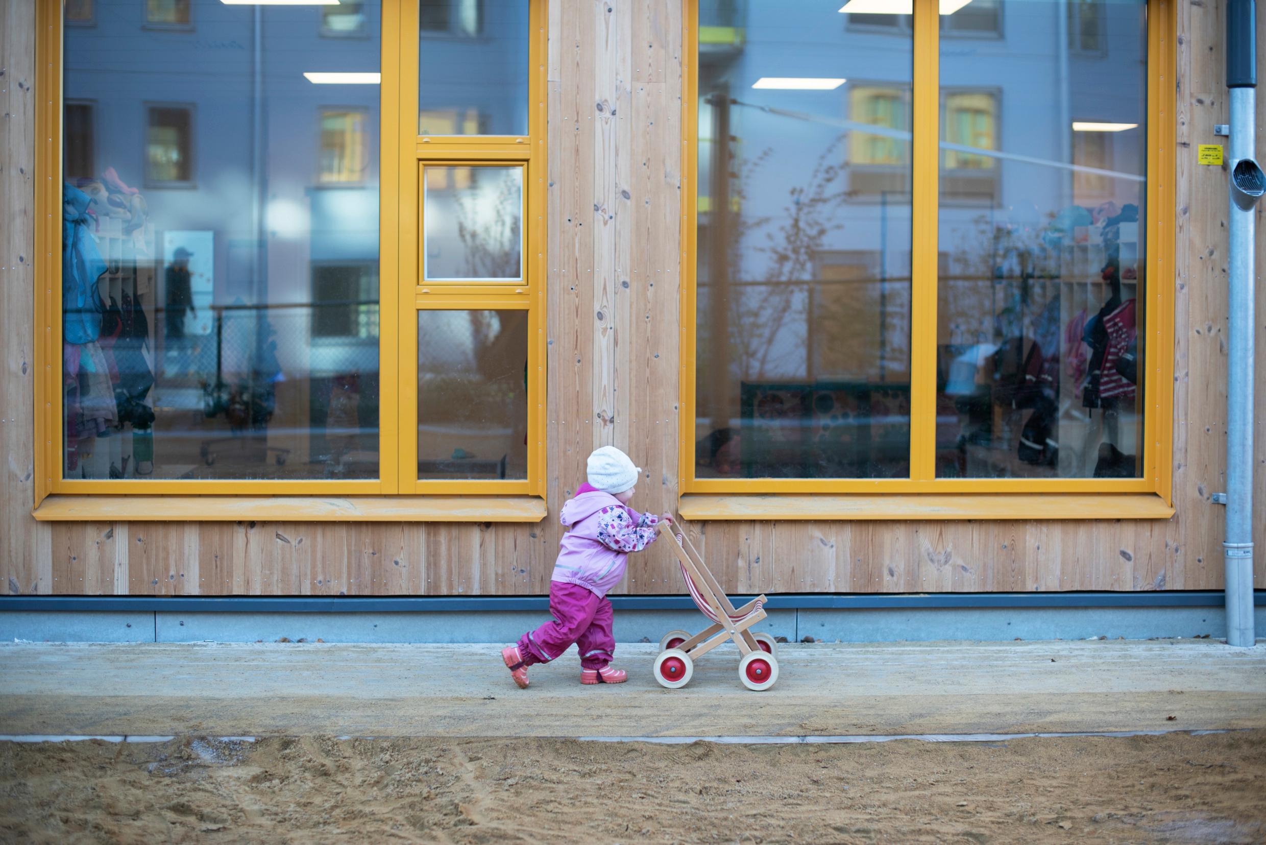 A toddler pushes a toy pram outside in front of a large window.