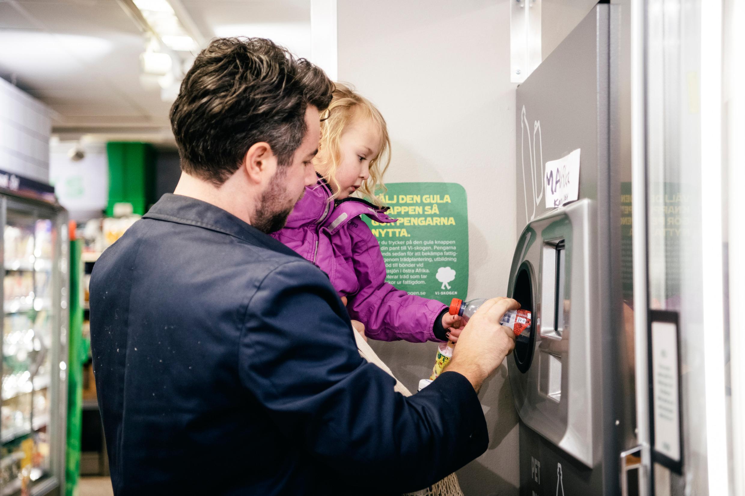 A man is holding a small child. Together they push a plastic bottle into a machine, A common sight of Swedish recycling.