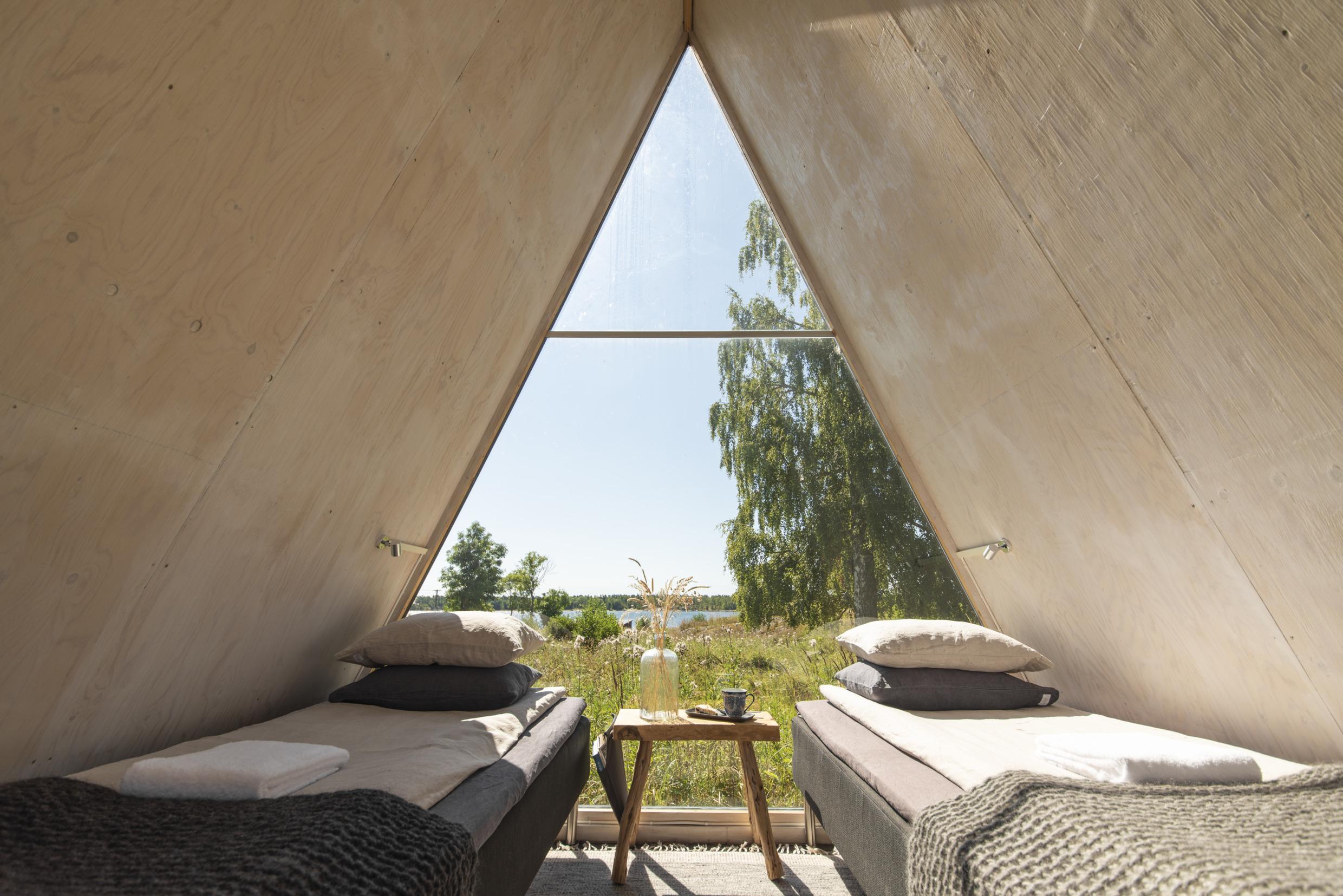 Interior view of modern beds and a bedside table inside a wooden a-frame cottage with triangular ceiling, looking out on a summer meadow of tall grass with the Baltic Sea behind it.