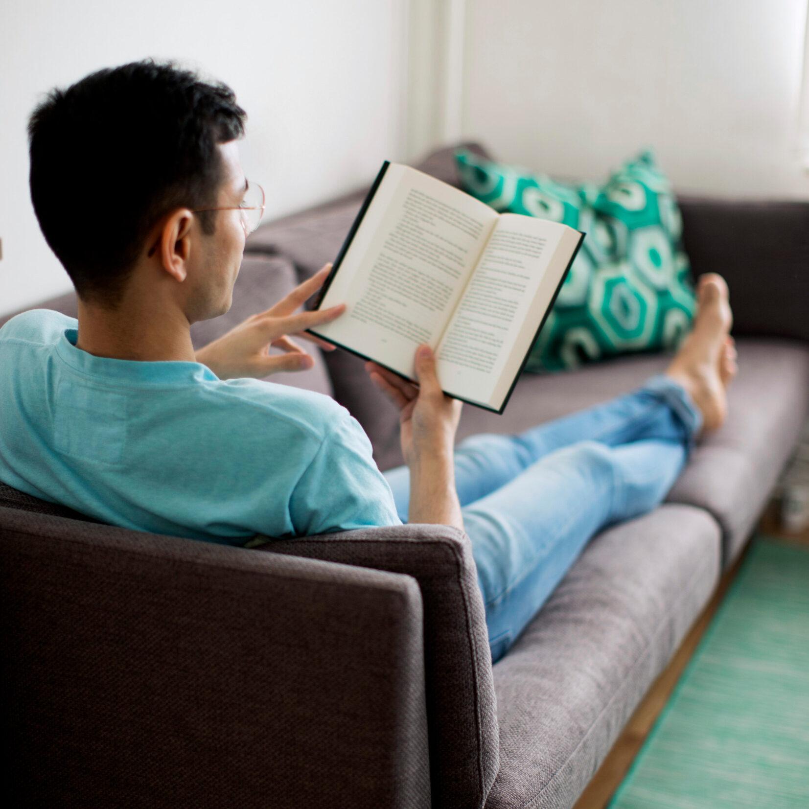 A man lounges in a sifa with a book in his hands.