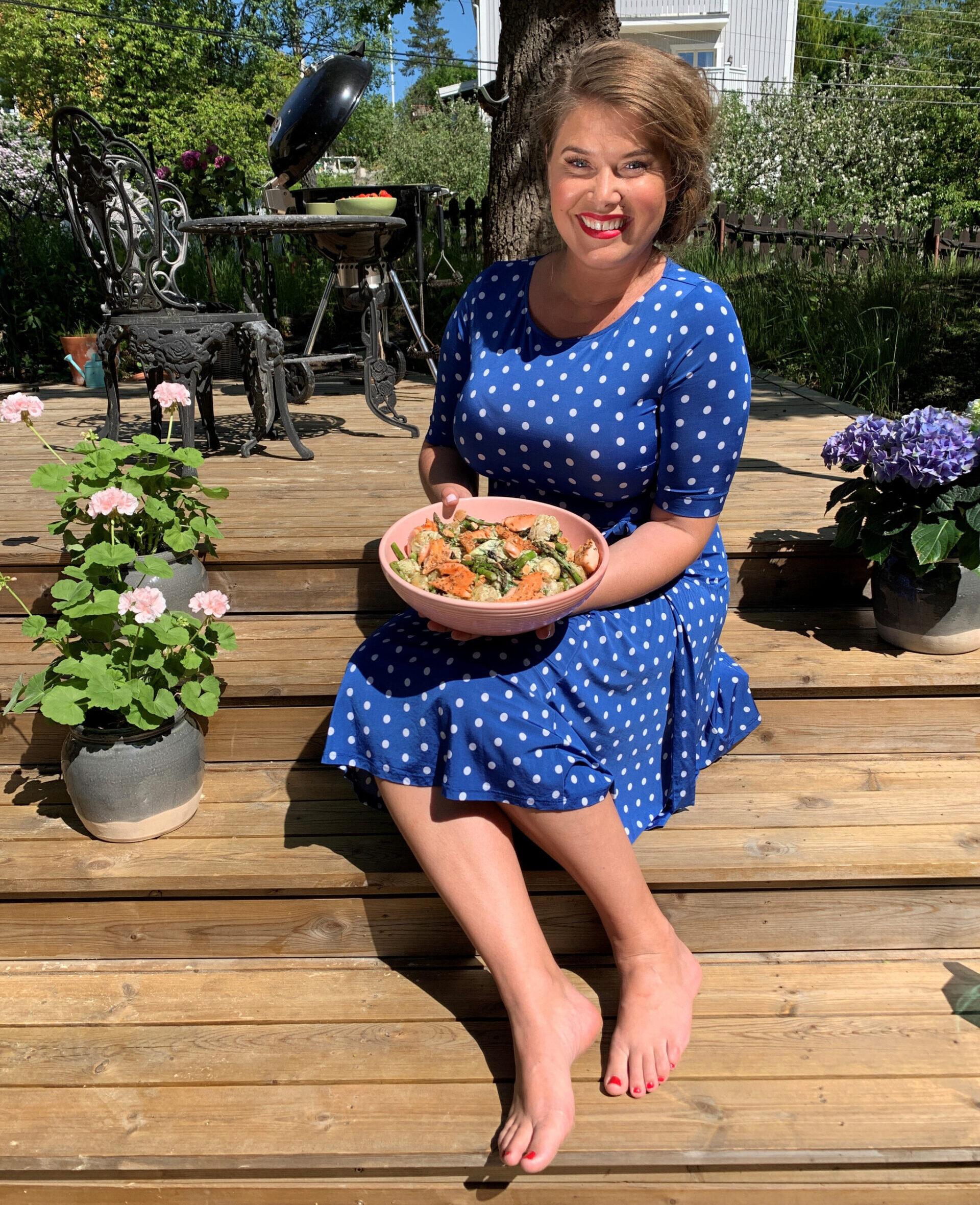 Jessica Frej sitting on some steps to an outdoor terrace, holding a bowl with food, flower pots next to her and a grill in the background.