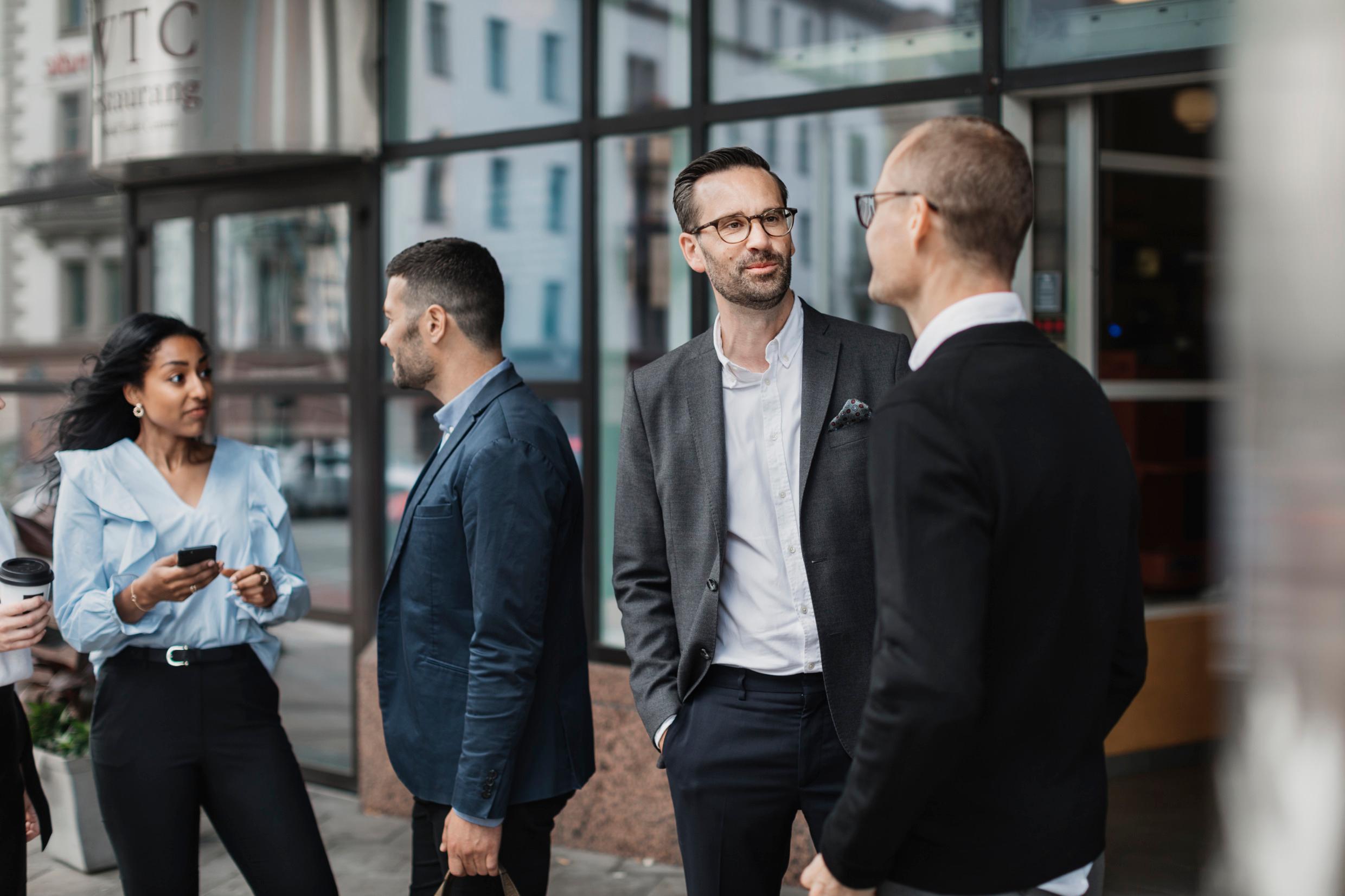 Outside a building are a group of people. Two men are talking to each other. A man and a woman talk to each other.