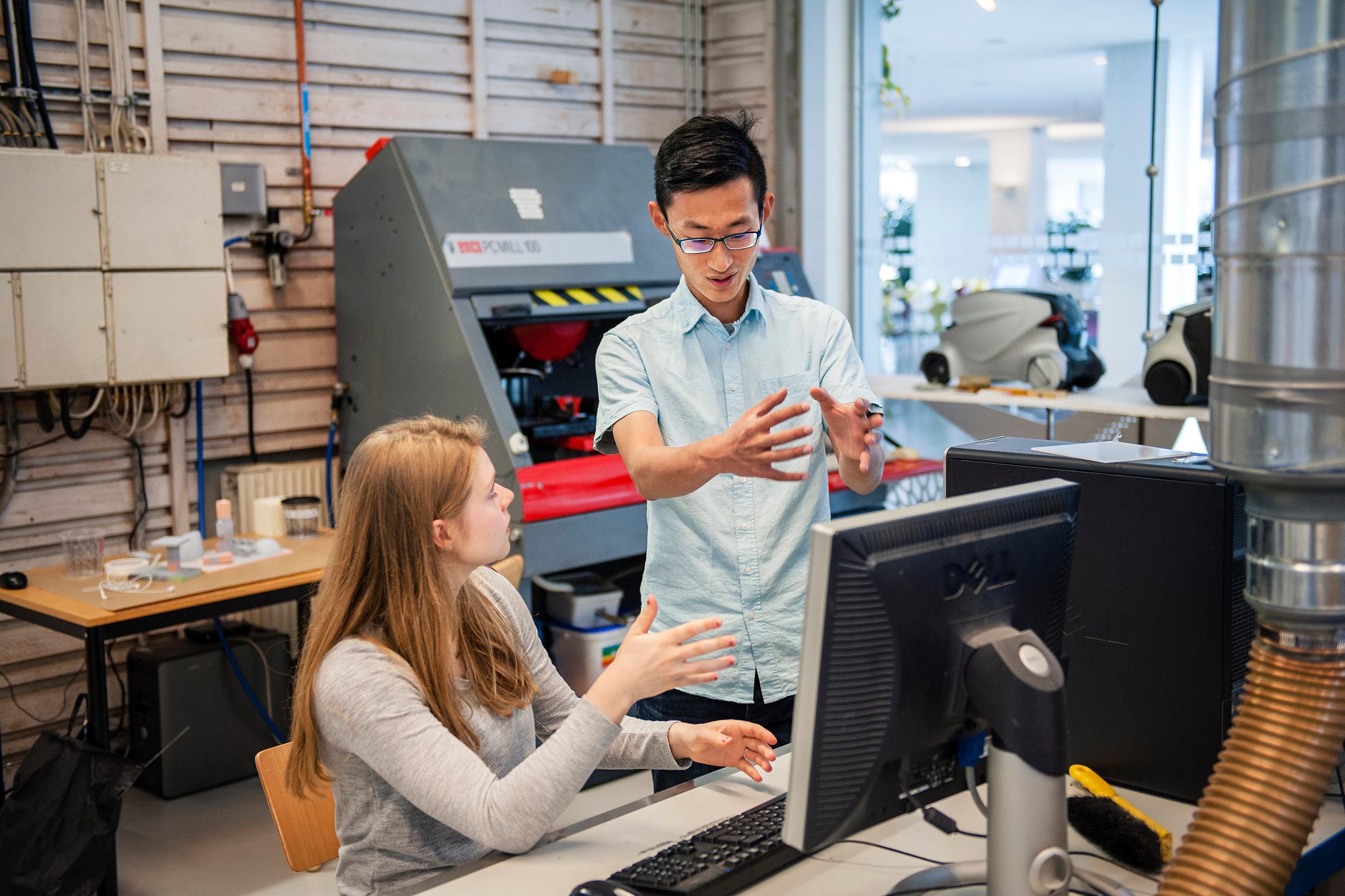 A woman sitting in front of a computer, her head turned away, and a man standing up next to her, showing something with his hands.