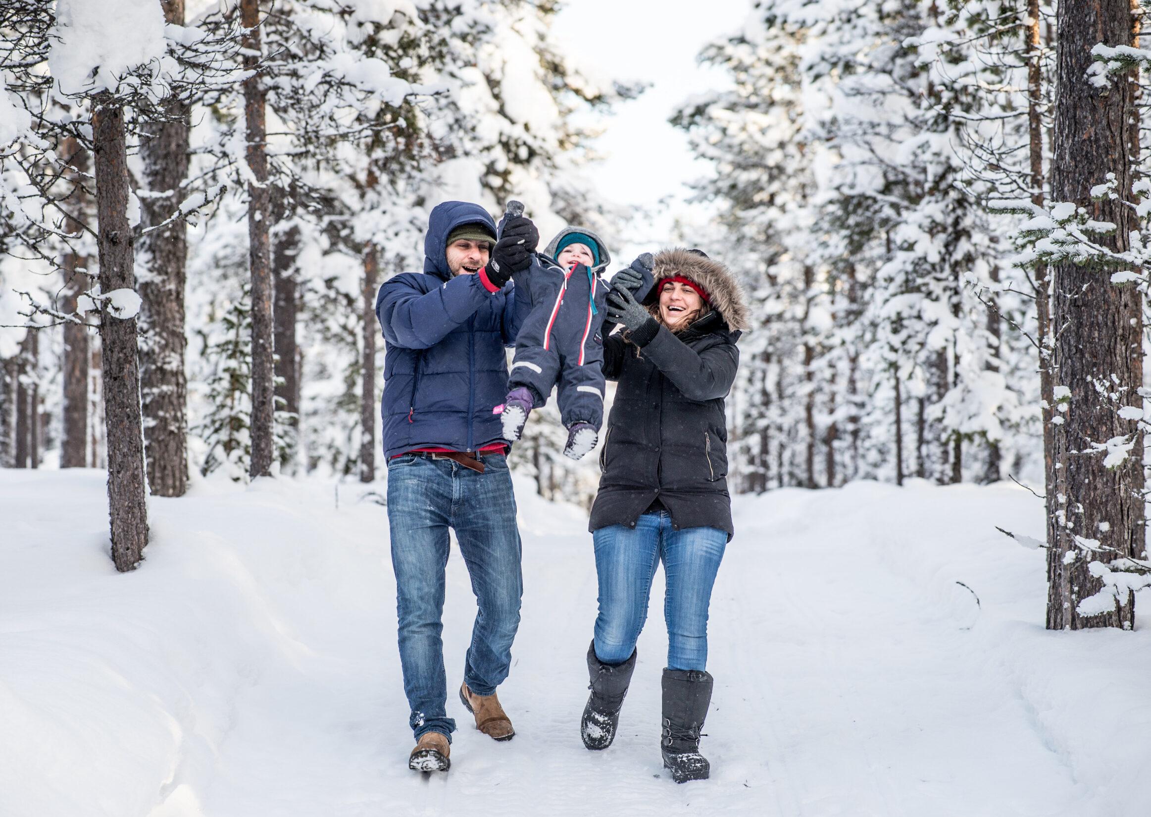 A man and a woman swings a toddler between them in a wintry forest landscape.