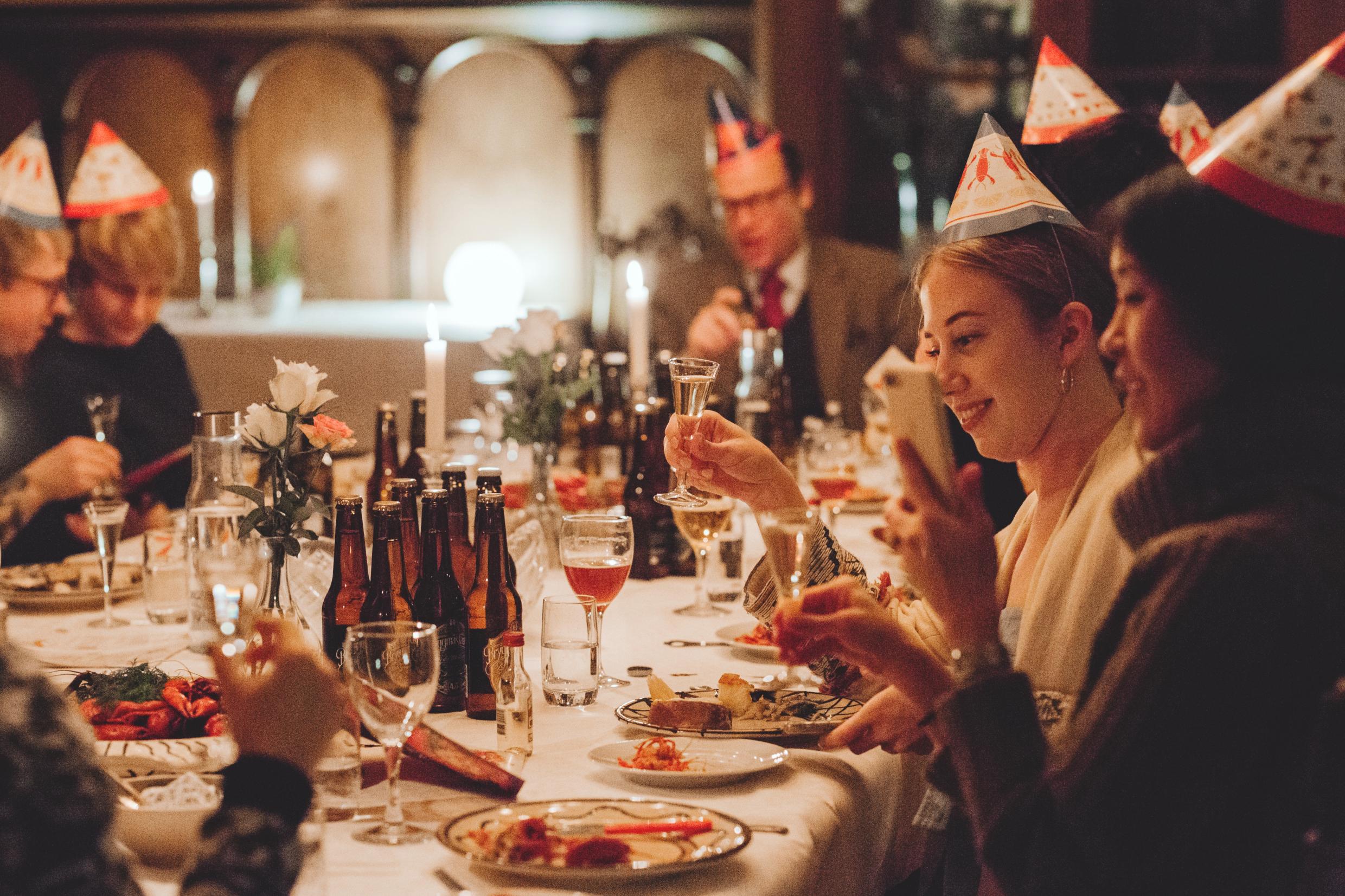 A group of people with party hats sitting around a table having crayfish and cheering with schnapps.