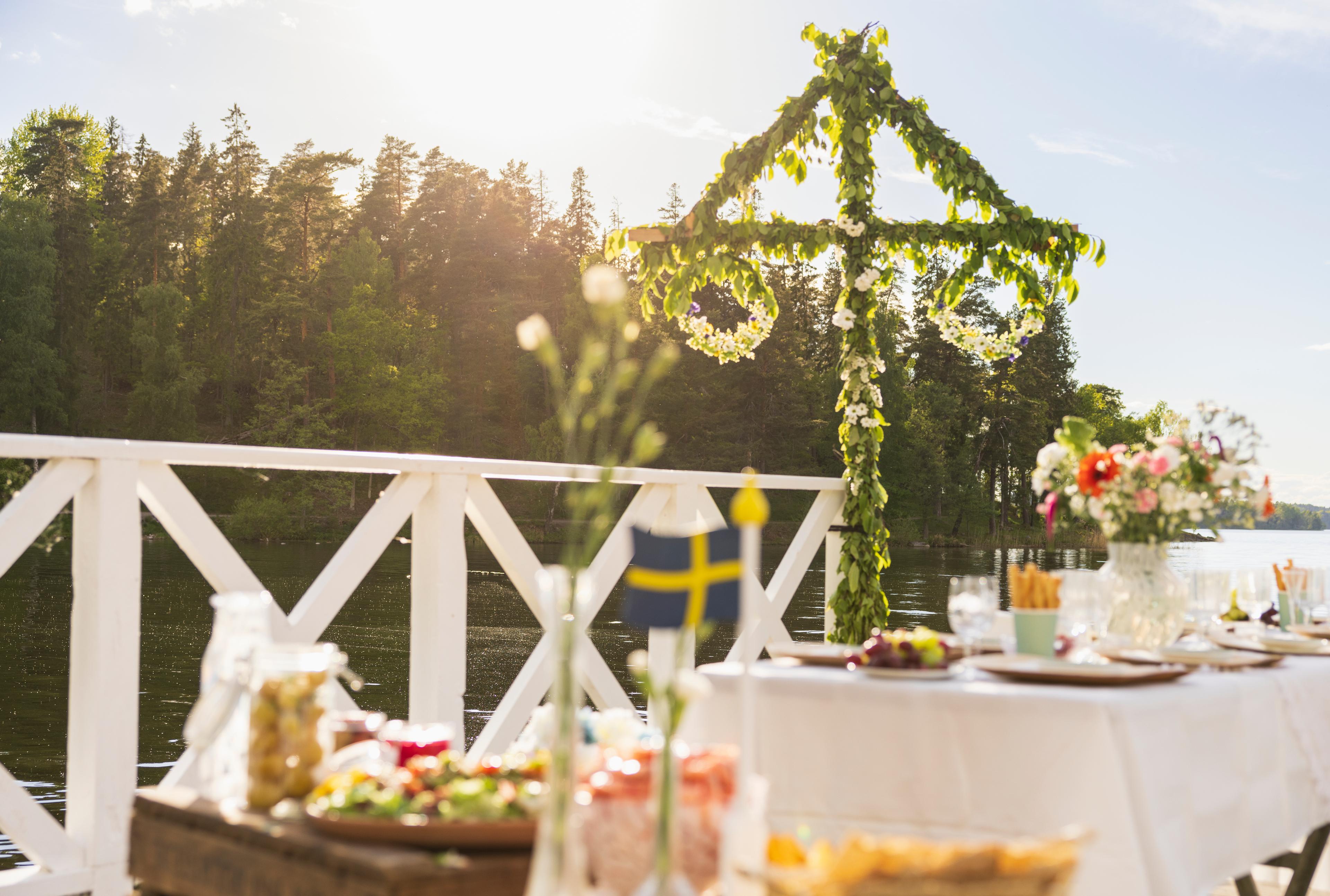 A festively set table on a pier with a Midsummer pole in the background