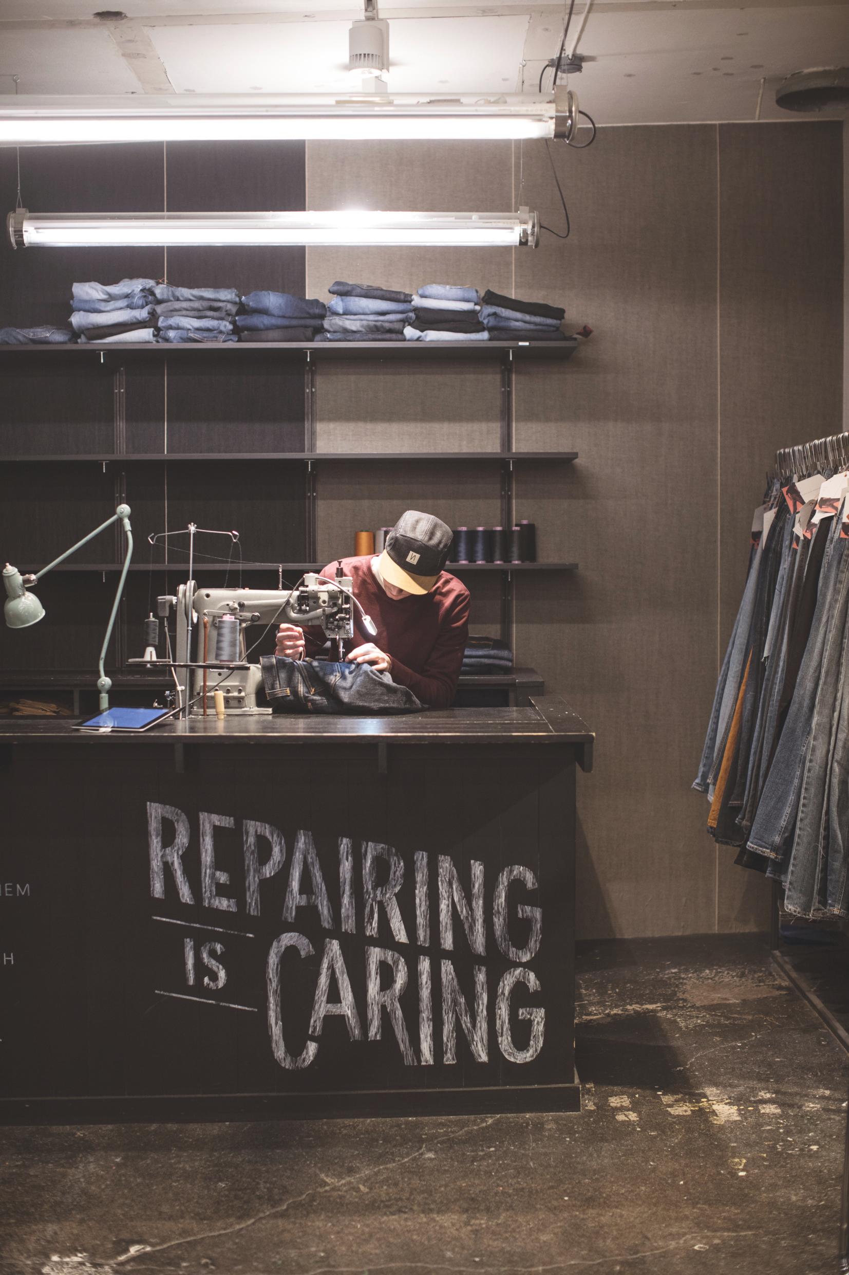 A man repairing a pair of jeans with a sewing machine, on a desk with a sign saying &quot;Repairing is caring&quot;.